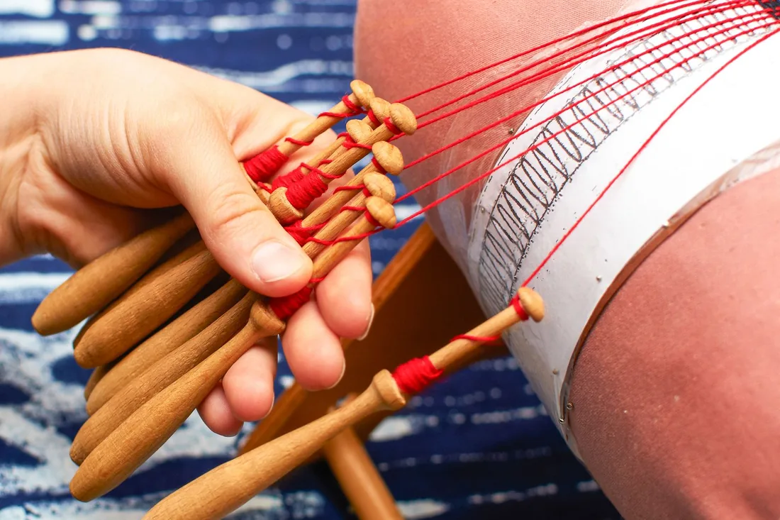 Manos sosteniendo varios bolillos de madera vestidos con hilo rojo sobre un mundillo cilíndrico con el picado alfilerado